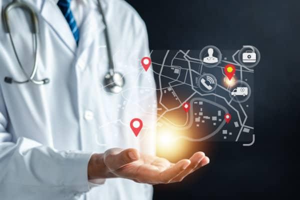 A doctor in a white coat and stethoscope holds out their hand, displaying a glowing digital map interface with red location pins and healthcare icons including an ambulance, medical kit, and person symbol.