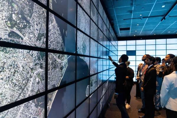 People observing a large wall of screens displaying maps and data in a control room or tech space.